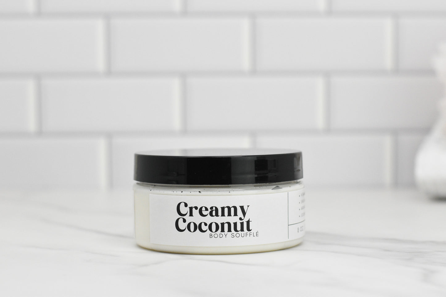 The image features a jar of 570 Soap Co.'s Creamy Coconut Body Butter, promising intense hydration. It is set on a marble surface, with the jar showcasing a black lid and a white label with black text, all against the backdrop of white subway tiles that evoke the feel of a tropical paradise.