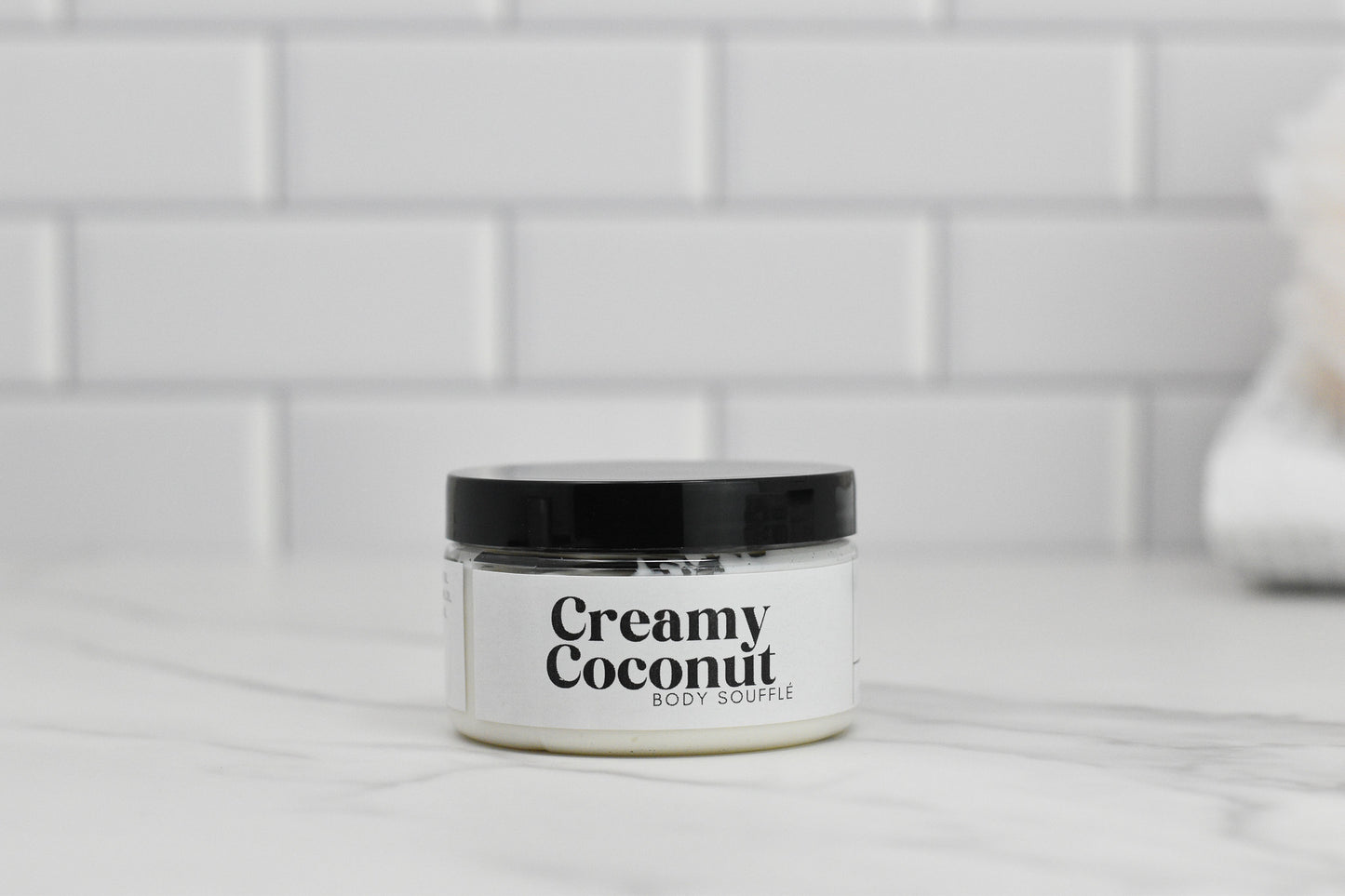 A jar of Creamy Coconut Body Butter from 570 Soap Co., adorned with a black lid, sits on a marble counter, promising intense hydration. The white tiled wall in the background evokes the feeling of a tropical paradise.