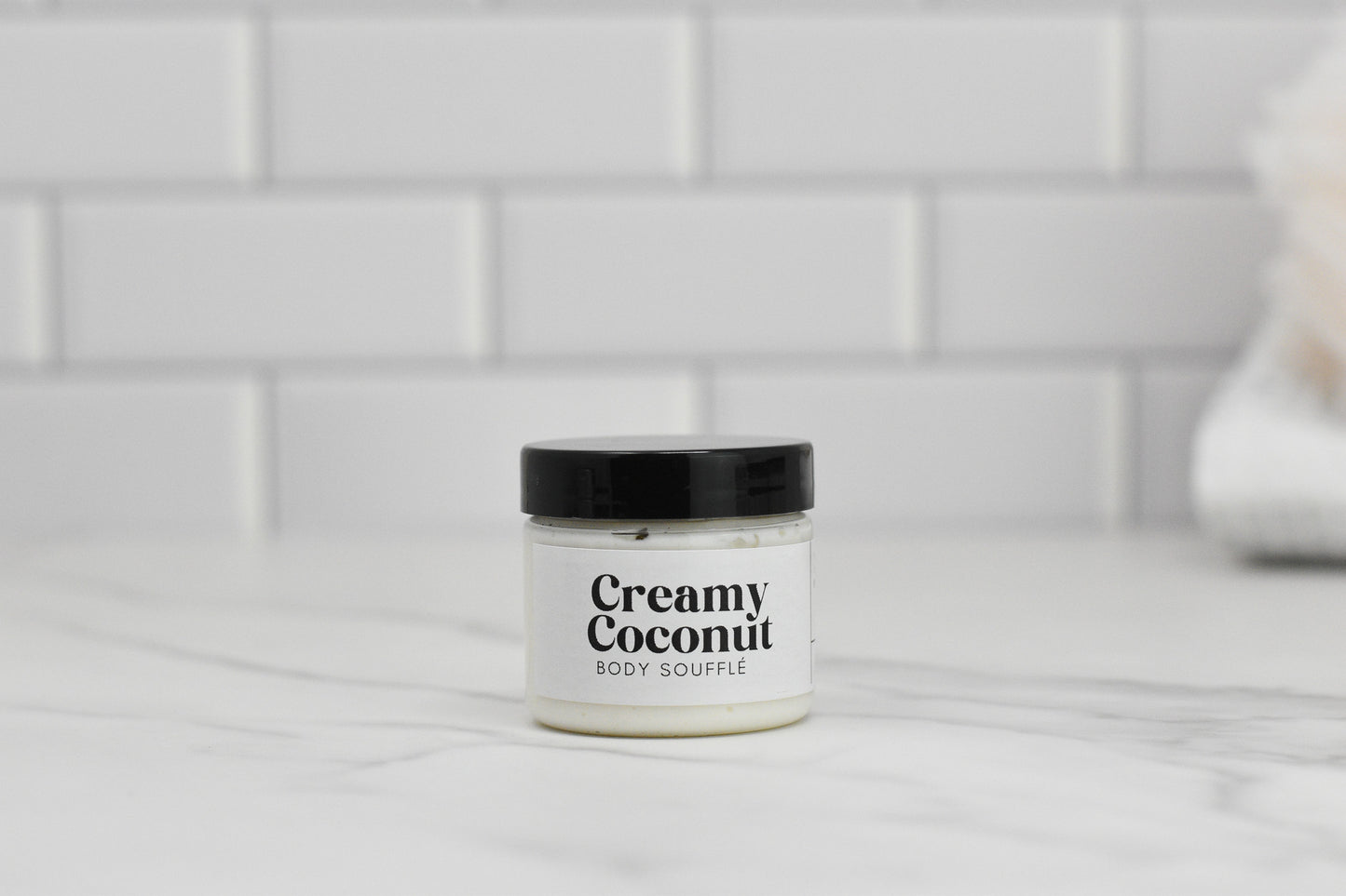 A jar of 570 Soap Co.’s Creamy Coconut Body Butter with a black lid is elegantly arranged on a marble surface against a backdrop of white tiles, offering intense hydration and capturing the essence of a tropical paradise.