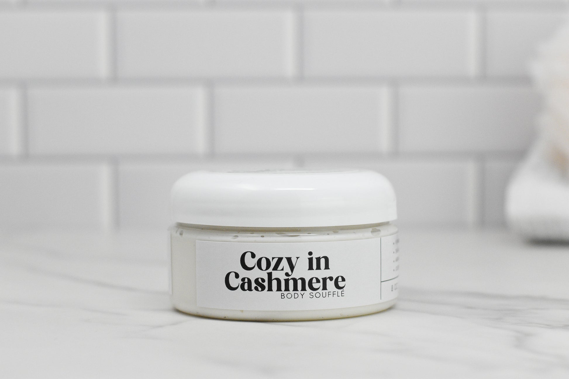 A jar of 570 Soap Co.'s Cozy in Cashmere Body Butter is elegantly placed on a marble surface with a white-tiled background, enriched with nourishing shea butter for ultimate hydration.