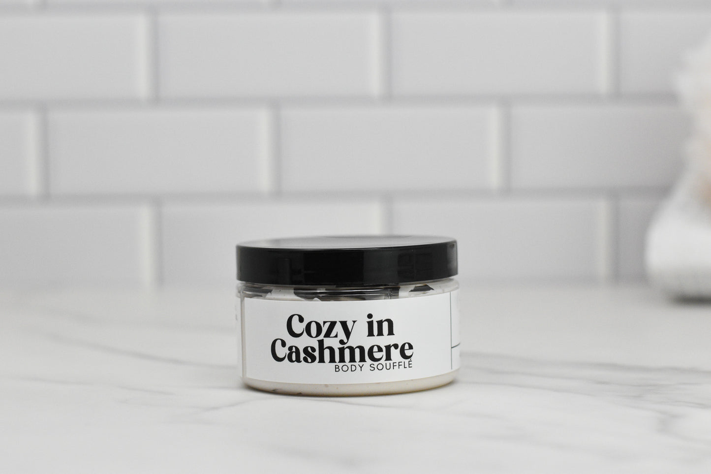 A jar of 570 Soap Co.'s Cozy in Cashmere Body Butter, featuring a black lid, sits on a marble countertop against a white tiled backsplash.