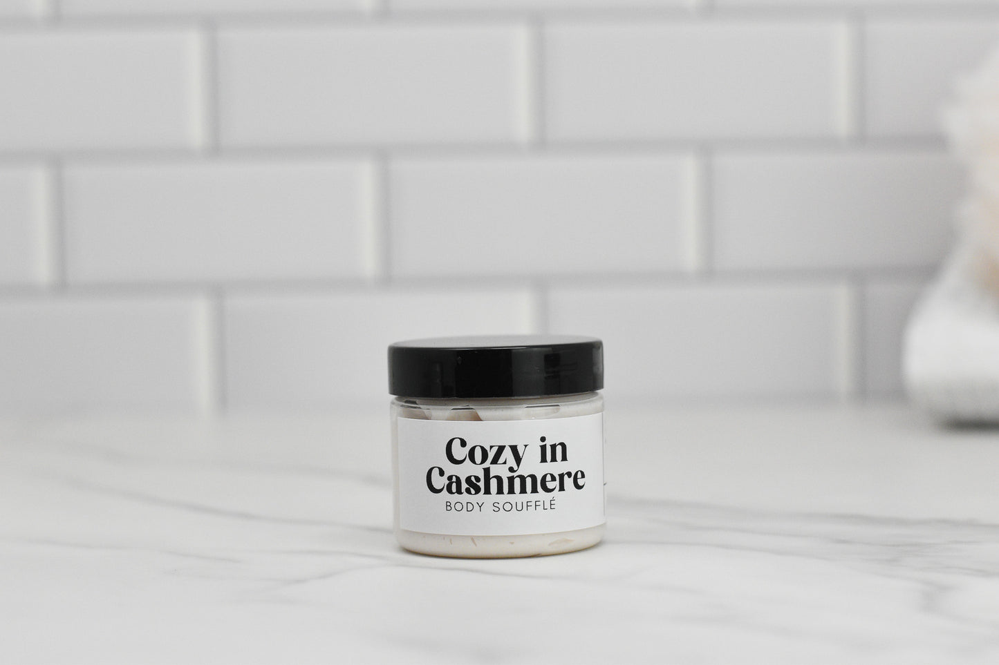 A jar of Cozy in Cashmere Body Butter by 570 Soap Co., enriched with shea butter and topped with a sleek black lid, sits elegantly on a white marble surface, while the serene backdrop of a white tile wall enhances its luxurious appeal.