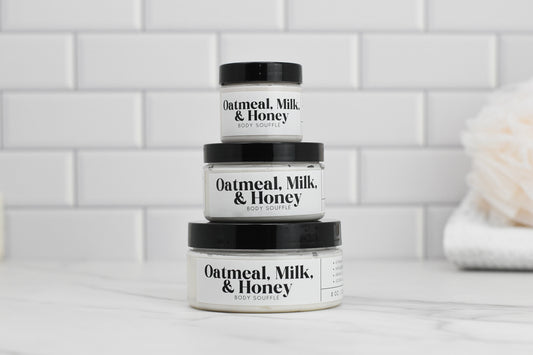 Three jars of Oatmeal Milk & Honey Body Butter by 570 Soap Co. infused with Shea Butter are stacked on a white tiled surface. The jars vary in size, arranged from the smallest on top to the largest on the bottom. A blurred towel is visible in the background.