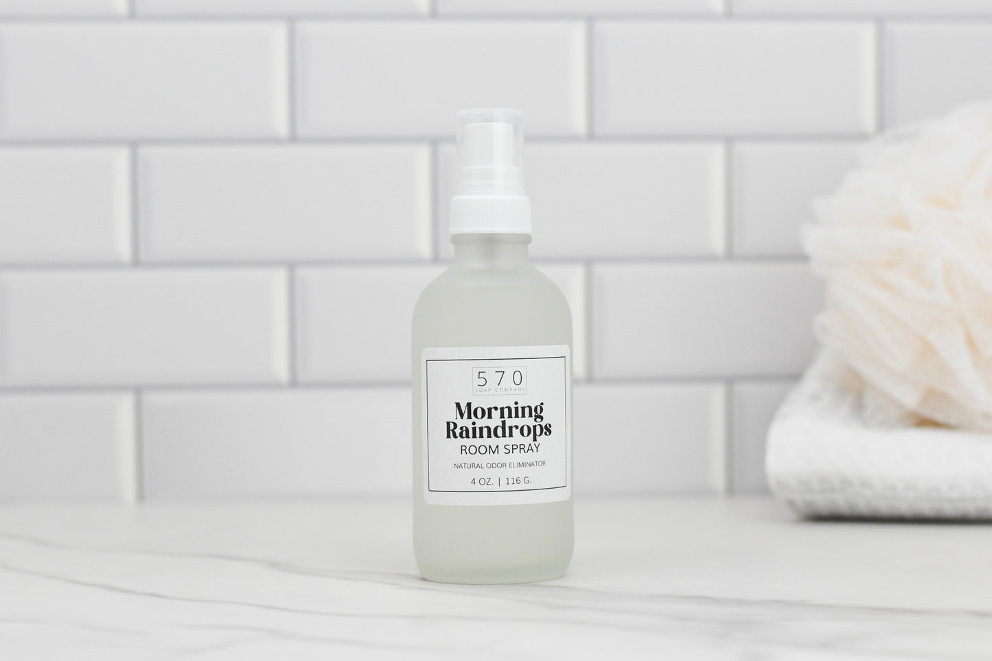 A frosted glass bottle labeled "570 Soap Co. Linen & Room Spray" with a white pump is placed on a marble surface. The background showcases white subway tiles and a blurred natural sponge to the right, emphasizing its function as a natural odor eliminator.