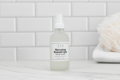 A frosted glass bottle labeled "570 Soap Co. Linen & Room Spray" with a white pump is placed on a marble surface. The background showcases white subway tiles and a blurred natural sponge to the right, emphasizing its function as a natural odor eliminator.