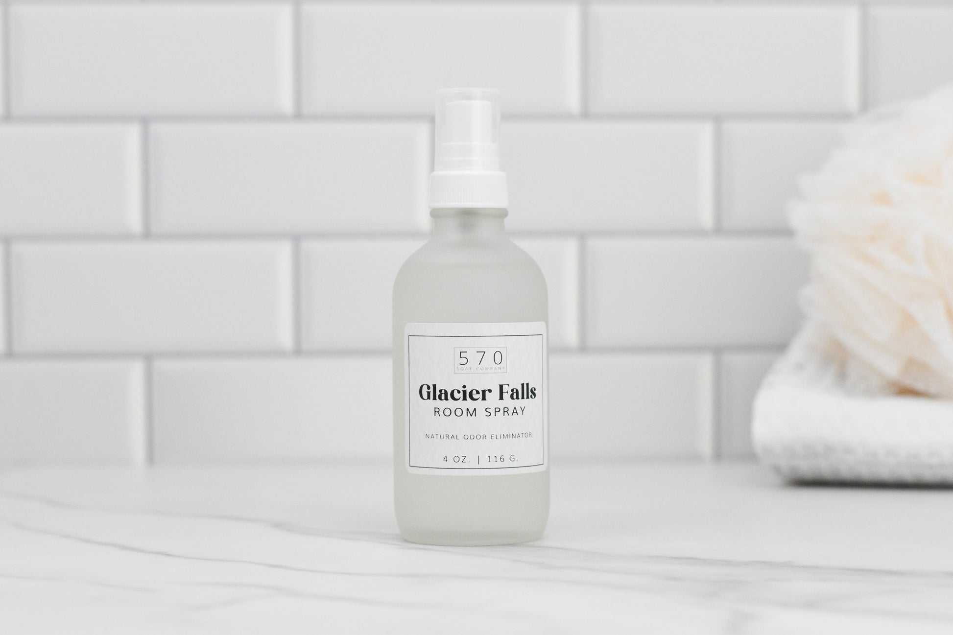 A bottle of 570 Soap Co. Linen & Room Spray is positioned on a marble surface. The label reads Glacier Falls, Natural Odor Eliminator, 4 oz | 116 ml. The background displays white subway tiles with a blurred towel and sponge to the right.