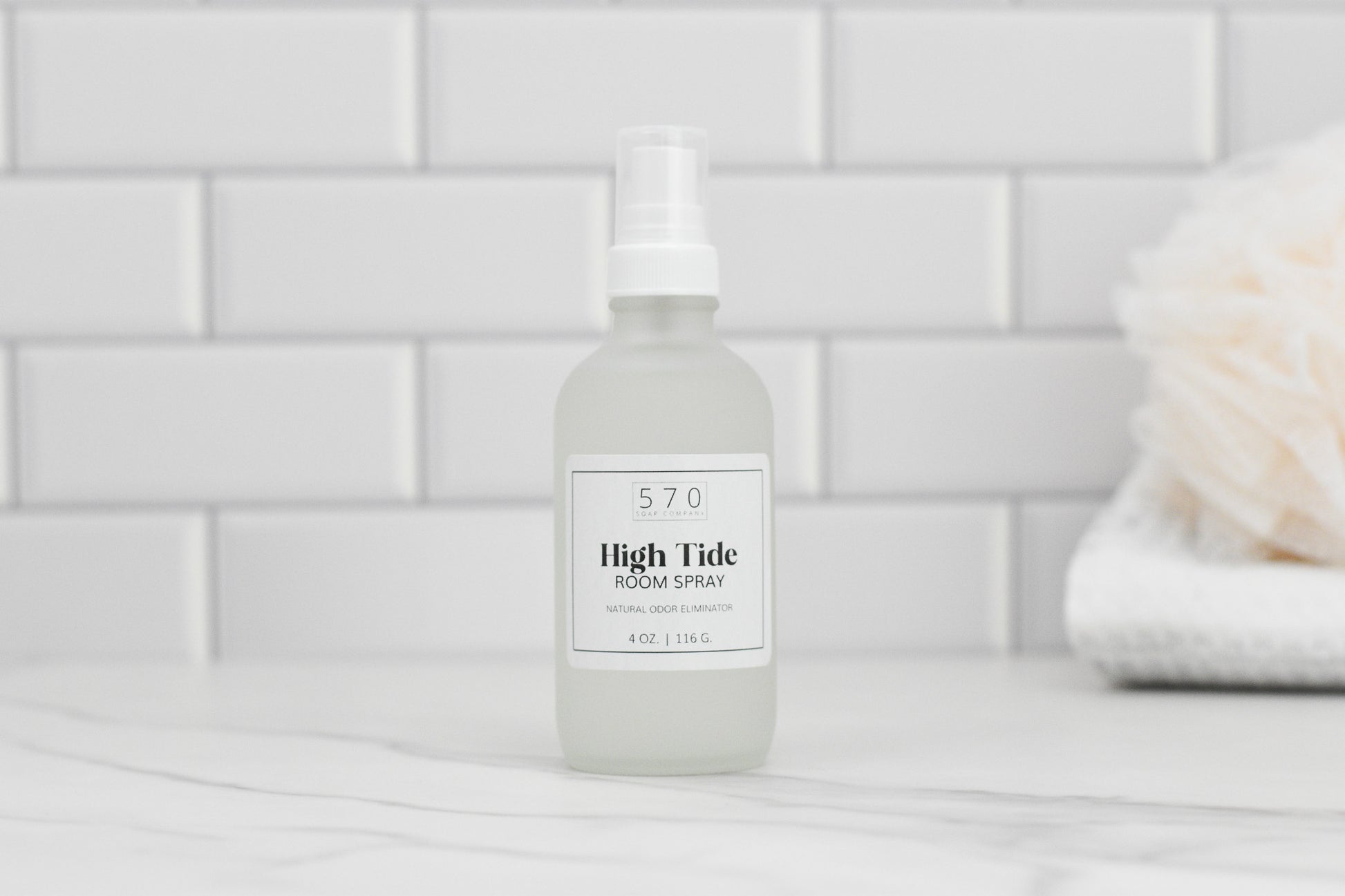 A frosted glass bottle of Linen & Room Spray by 570 Soap Co., featuring a white label, is placed on a marble surface. The label indicates 4 oz | 118 g High Tide Room Spray. Positioned against a white tiled background, it sits next to a soft bath sponge. Ideal for use as both linen and room spray, offering various fragrances.