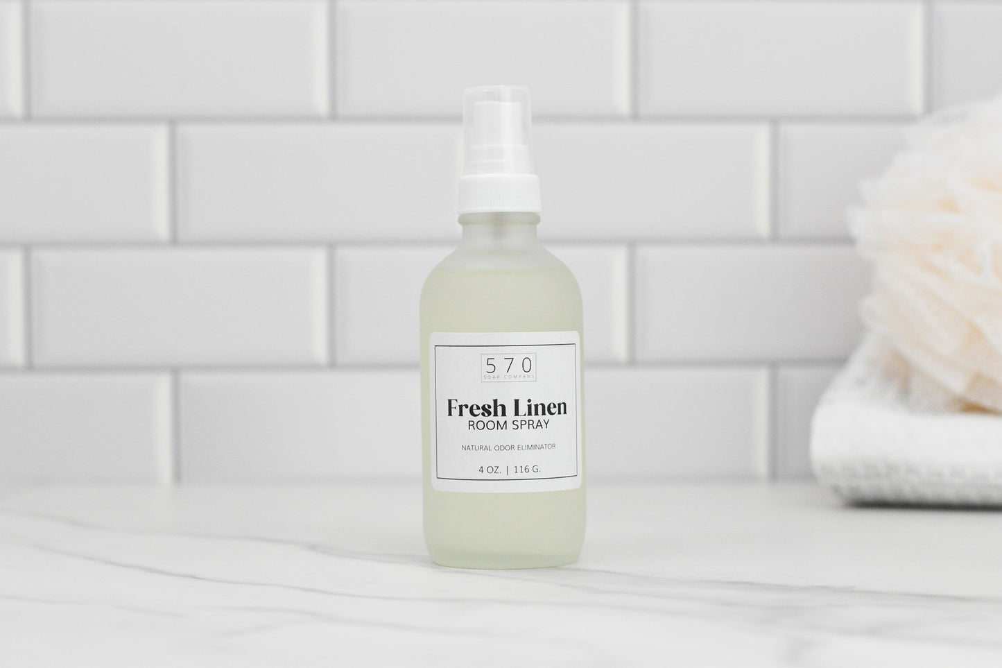 A bottle of 570 Soap Co.'s Linen & Room Spray sits on a white countertop with a tiled backsplash in view. Adorned with a label and spray nozzle, this natural odor eliminator refreshes the air effortlessly.