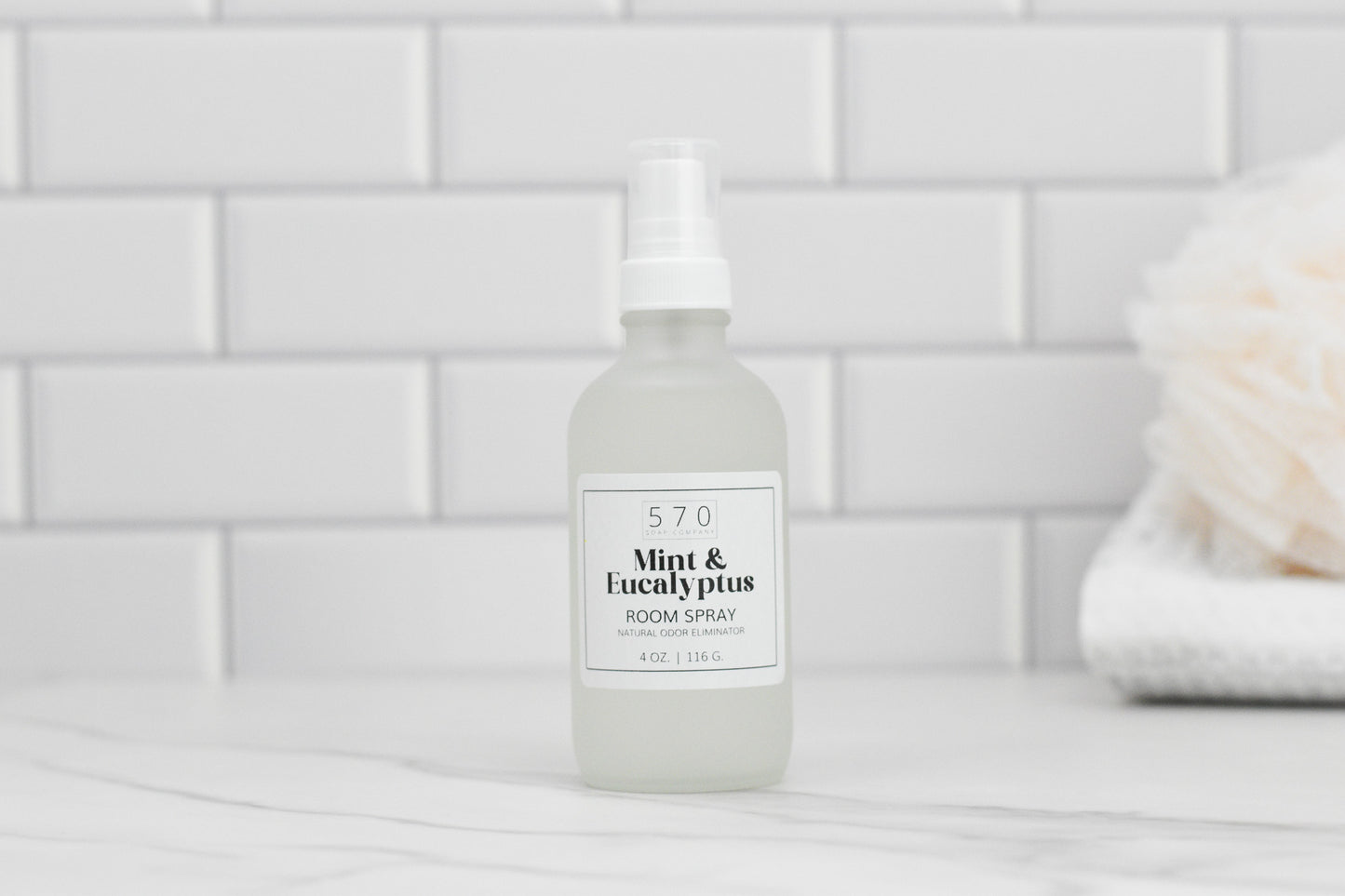 A bottle of 570 Soap Co.'s Linen & Room Spray, celebrated for its natural odor-eliminating properties, is labeled Mint & Eucalyptus and sits on a marble surface. The background highlights white subway tiles with a blurred soft object on the right, hinting at its dual-purpose use as both a linen and room spray.