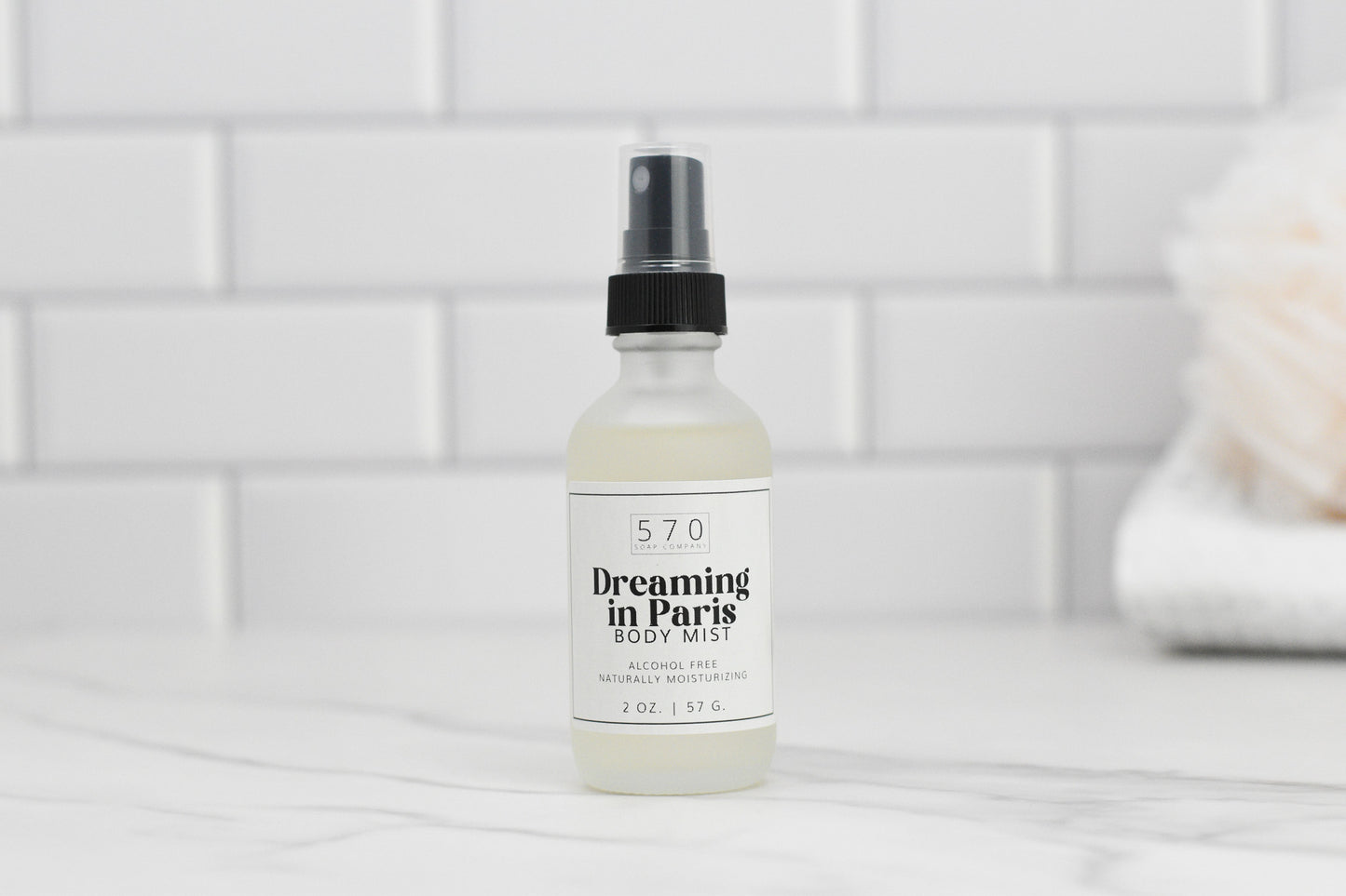 A bottle of 570 Soap Co.'s Luxurious Fragranced Body Mist Spray rests on a marble surface. Renowned for its alcohol-free formula and advanced hydrating technology, it stands before a white tiled backsplash, adding a clean and modern touch to the image.