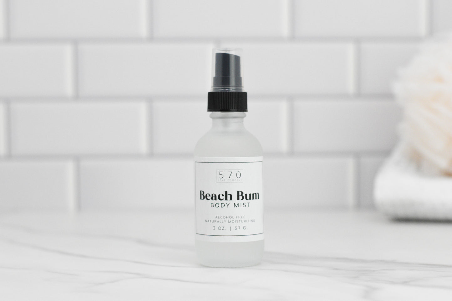 A frosted glass bottle of Luxurious Fragranced Body Mist Spray by 570 Soap Co., featuring hydrating technology and a black spray nozzle, sits on a white marble surface. The tiled white background and a soft bath sponge contribute to a clean, spa-like atmosphere. The label highlights its alcohol-free and moisturizing properties.