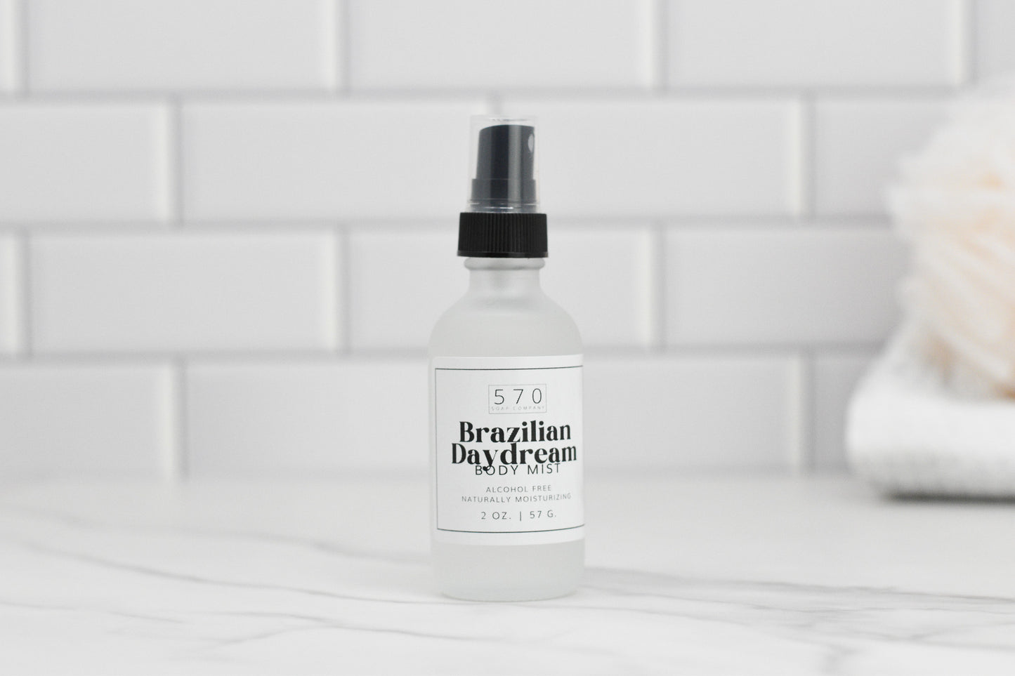 A clear bottle of Luxurious Fragranced Body Mist Spray by 570 Soap Co., featuring a black spray top, sits on a marble surface, proudly showcasing its alcohol-free formula. The label reveals it contains 2 oz. of product. White subway tiles and a blurred object add to the serene background ambiance.