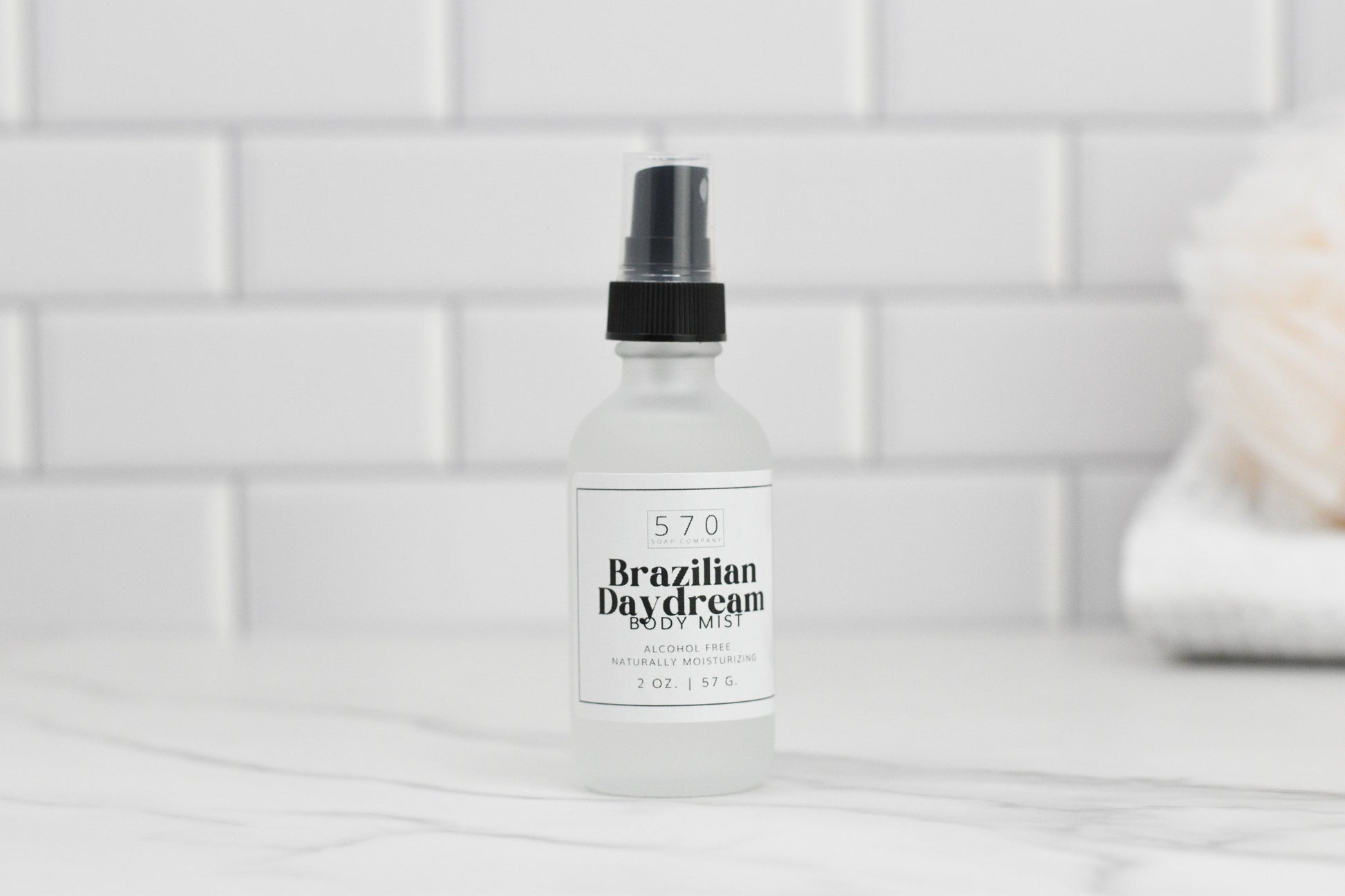 A clear bottle of Luxurious Fragranced Body Mist Spray by 570 Soap Co., featuring a black spray top, sits on a marble surface, proudly showcasing its alcohol-free formula. The label reveals it contains 2 oz. of product. White subway tiles and a blurred object add to the serene background ambiance.