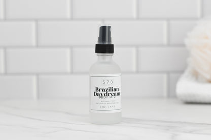 A clear bottle of Luxurious Fragranced Body Mist Spray by 570 Soap Co., featuring a black spray top, sits on a marble surface, proudly showcasing its alcohol-free formula. The label reveals it contains 2 oz. of product. White subway tiles and a blurred object add to the serene background ambiance.