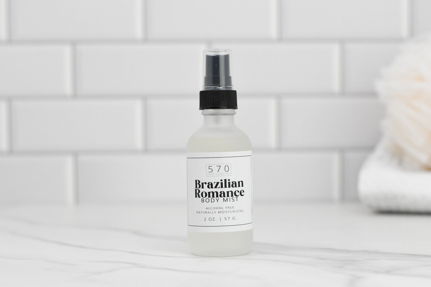 A bottle of Luxurious Fragranced Body Mist Spray by 570 Soap Co. rests on a white tiled surface. The alcohol-free label emphasizes its innovative hydrating technology, featuring a 2 oz (57 g) capacity. A soft, blurred background highlights the clean and minimalist aesthetic.