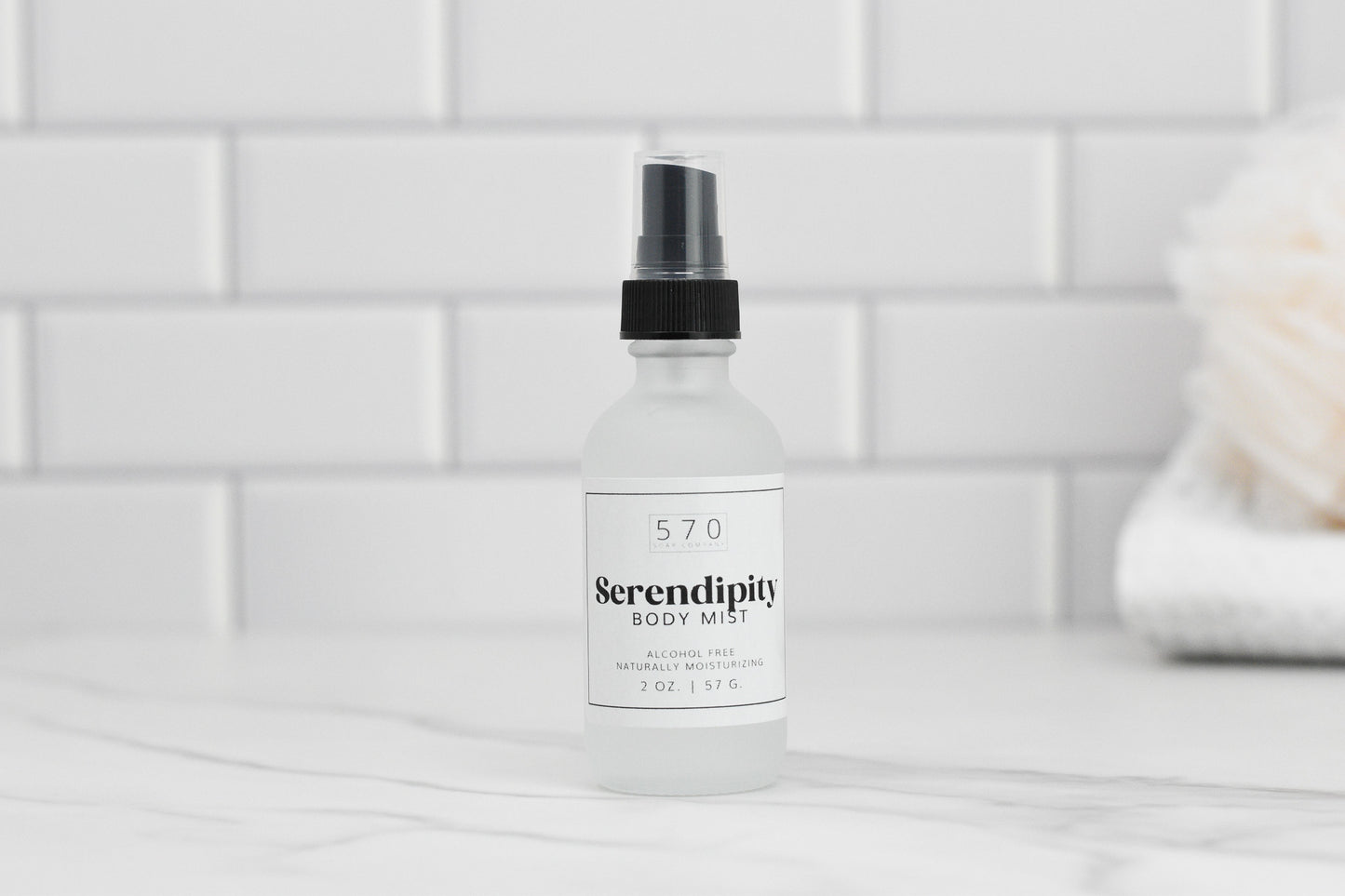 A bottle of Luxurious Fragranced Body Mist Spray by 570 Soap Co., designed with hydrating technology and a convenient spray nozzle, is labeled as alcohol-free and naturally moisturizing. This 2 oz (57 g) bottle is elegantly placed on a white marble surface against a backdrop of pristine white tiles.