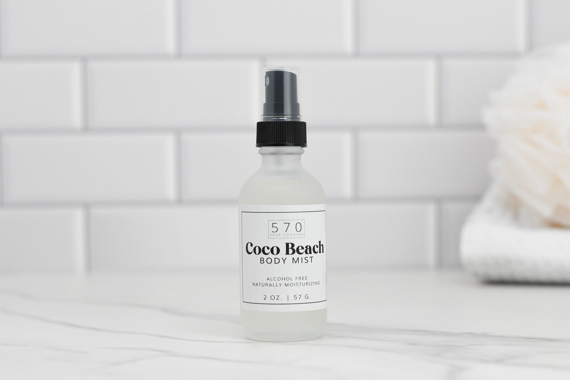 A frosted glass bottle labeled "Luxurious Fragranced Body Mist Spray" by 570 Soap Co. with a black spray cap rests on a white marble surface, highlighting its alcohol-free formula. The backdrop includes white subway tiles and a blurred white towel paired with a beige loofah, enhancing the scene's refreshing ambiance.