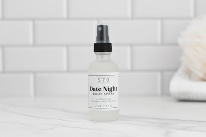 A bottle of Luxurious Fragranced Body Mist Spray from 570 Soap Co., highlighting its hydrating technology and naturally moisturizing formula, rests on a marble countertop. The background features white subway tiles with a soft-focus object on the right.