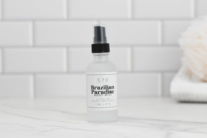 A bottle of 570 Soap Co.'s Luxurious Fragranced Body Mist Spray, boasting advanced hydrating technology and labeled as naturally moisturizing, sits on a white tiled counter. It contains 2 ounces (57 grams) of product. A fluffy white object is blurred in the background.