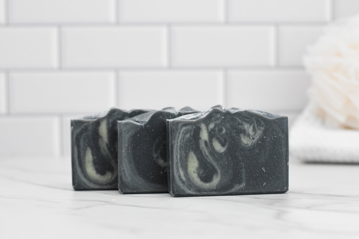 Three bars of 570 Soap Co.'s Tea Tree & Charcoal Bar Soap – All Natural are elegantly arranged on a marble surface, set against a backdrop of white tiled walls.