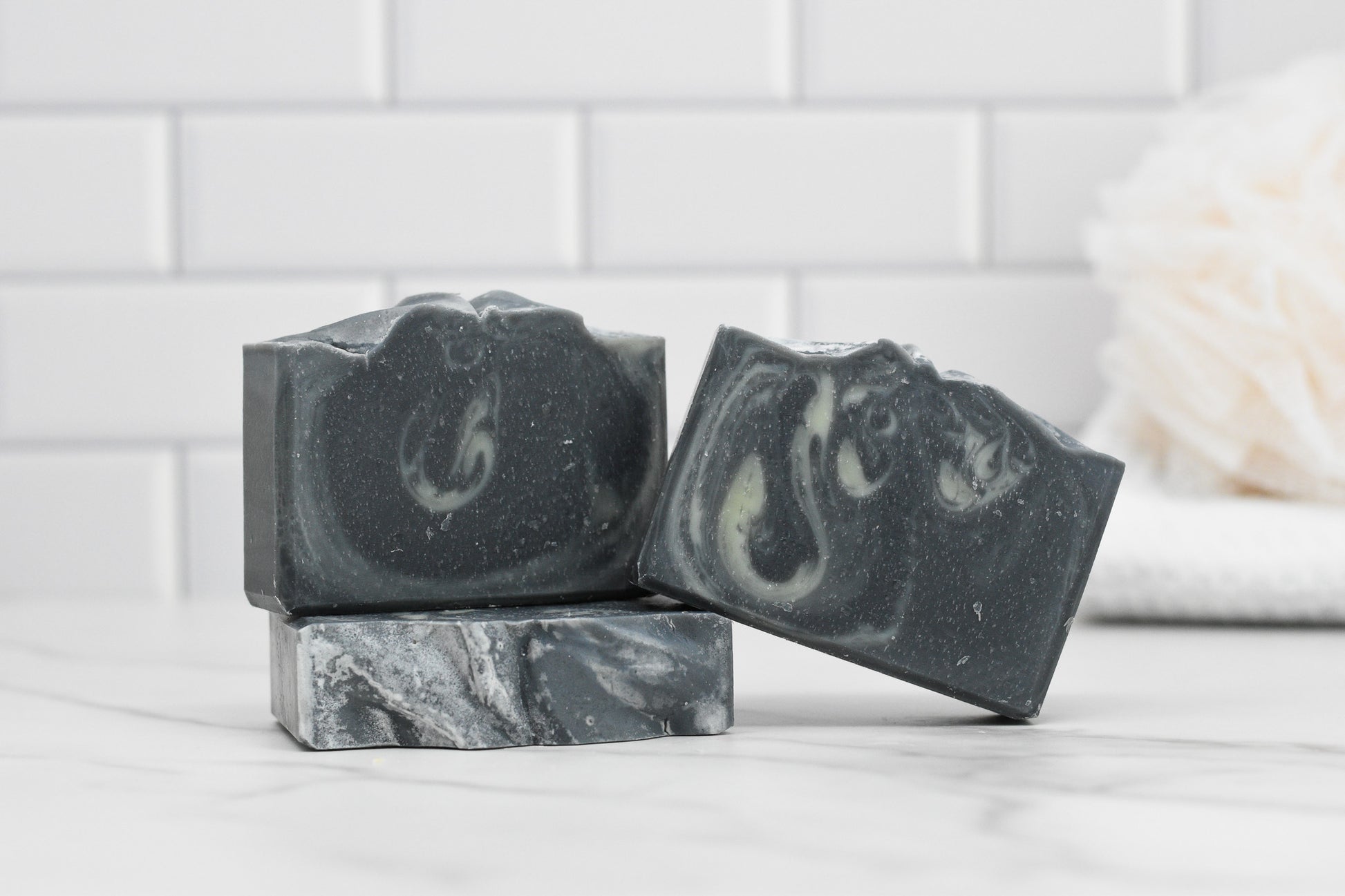 Three bars of 570 Soap Co.'s Tea Tree & Charcoal Bar Soap - All Natural, featuring a dark gray color with white swirls, are stacked on a marble countertop. Infused with activated charcoal and tea tree essential oil, they rest against a backdrop of white subway tiles alongside a soft white bath sponge.