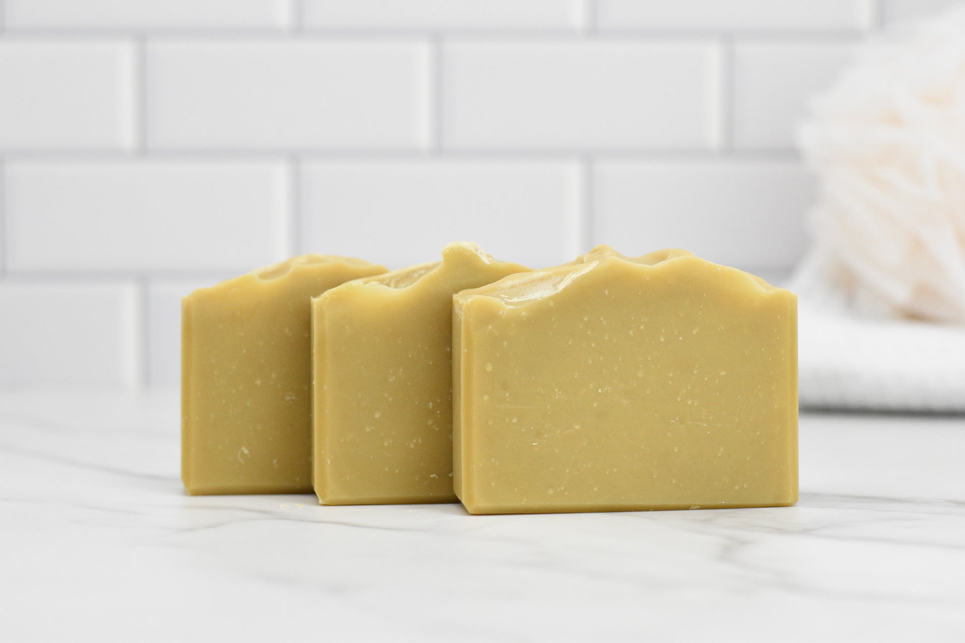 Three Sun-Kissed Lemonade Bar Soaps by 570 Soap Co., in a vibrant olive green and infused with a refreshing citrus zest, are neatly arranged on a marble surface. The background showcases sleek white subway tiles and a softly blurred bath sponge in the upper right corner, adding an elegant touch to the soothing scene.