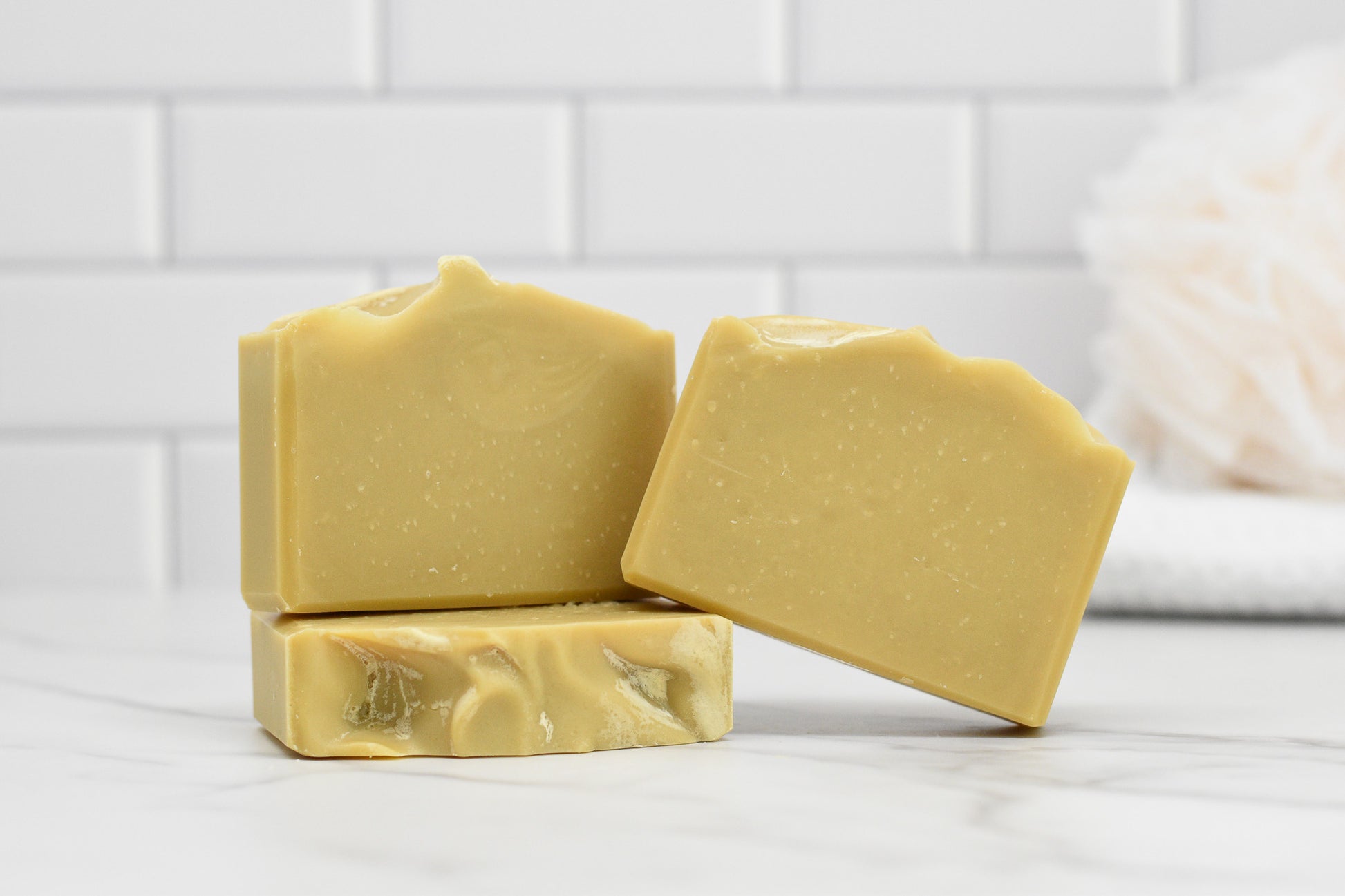 Three bars of Sun-Kissed Lemonade Bar Soap by 570 Soap Co., boasting a smooth, creamy texture, are stacked on a white marble surface. The background features white subway tiles and a softly blurred bath sponge, hinting at the citrusy zest within.