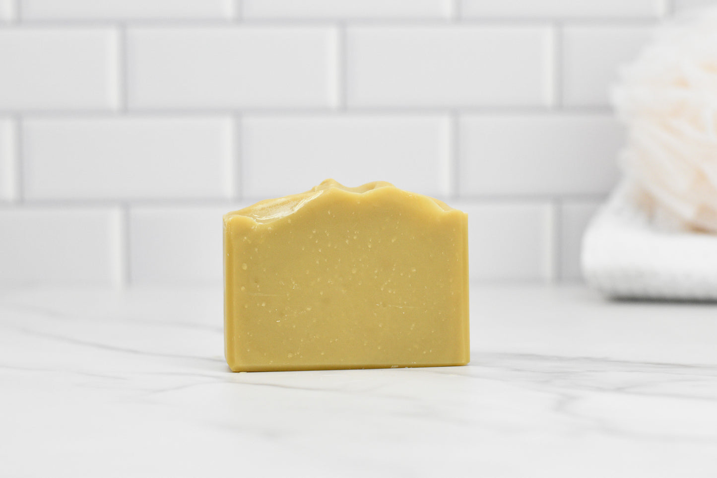 A Sun-Kissed Lemonade Bar Soap by 570 Soap Co., featuring a handcrafted wavy top, rests on a white marble surface. The background showcases white subway tiles, and a soft towel is partially visible on the right side, infusing the serene setting with refreshing citrusy zest.