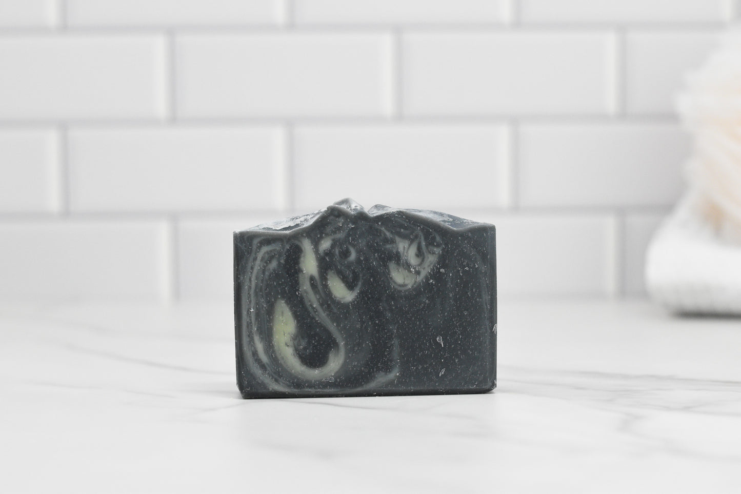 A bar of Tea Tree & Charcoal Bar Soap - All Natural by 570 Soap Co. with white marbled swirls rests on a marble countertop. The background is adorned with white subway tiles, and a towel is slightly blurred on the right edge.