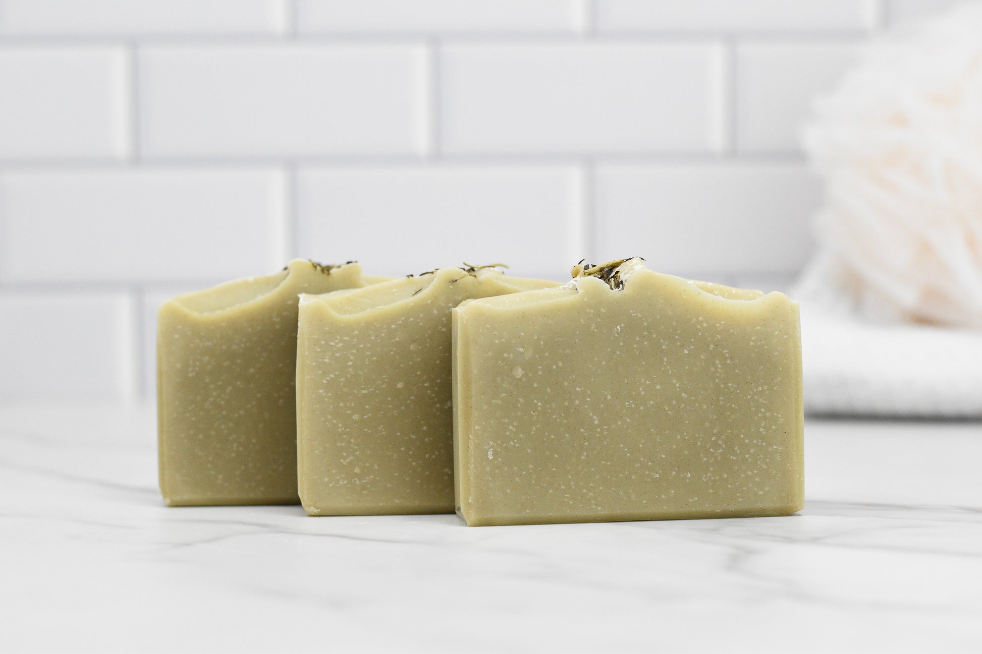 Three bars of 570 Soap Co.'s Rosemary Peppermint Bar Soap - All Natural are neatly arranged on a marble countertop. The background features a white subway tile wall and a blurred white bath sponge, creating a clean, minimalist aesthetic that emphasizes natural ingredients and gentle exfoliation.