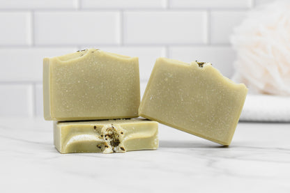 Three bars of 570 Soap Co.'s Rosemary Peppermint Bar Soap rest elegantly on a marble surface. Each beige handmade soap features a speckled texture, with one bar infused with rosemary peppermint essential oil and adorned with dried herbs. The backdrop of white subway tiles and the blurred silhouette of a white loofah accentuates the soap's all-natural ingredients.