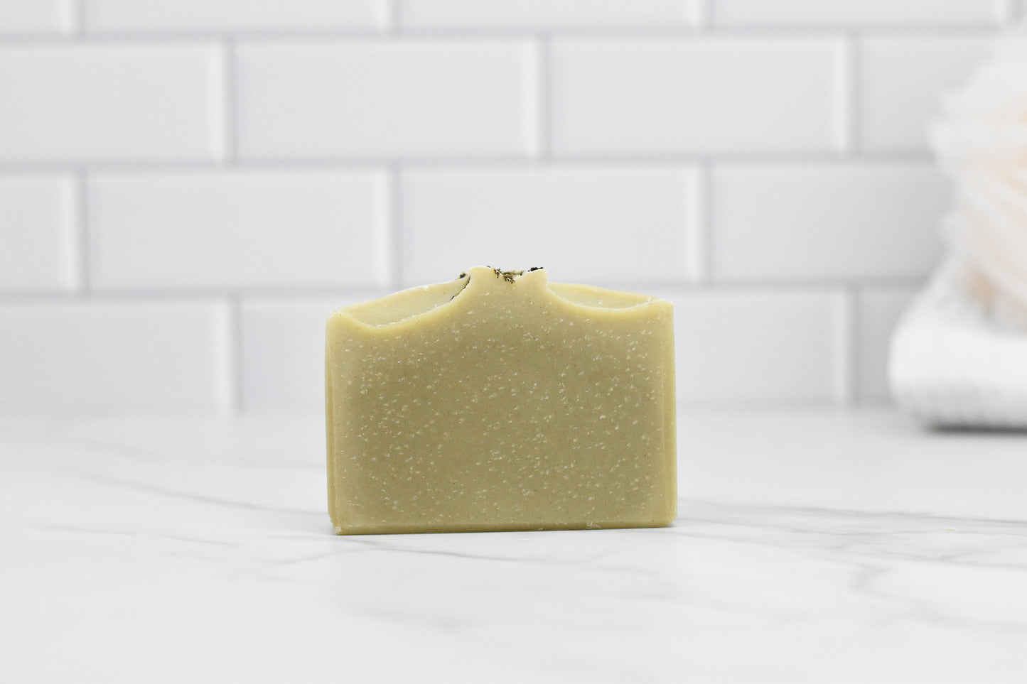 A bar of 570 Soap Co.'s Rosemary Peppermint Bar Soap - All Natural rests on a smooth, white marble surface. Its light, natural hue and delicate texture suggest gentle exfoliation. In the background, a soft-focus white tile wall provides contrast to a folded towel visible on the right.