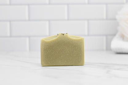 A bar of 570 Soap Co.'s Rosemary Peppermint Bar Soap - All Natural rests on a smooth, white marble surface. Its light, natural hue and delicate texture suggest gentle exfoliation. In the background, a soft-focus white tile wall provides contrast to a folded towel visible on the right.