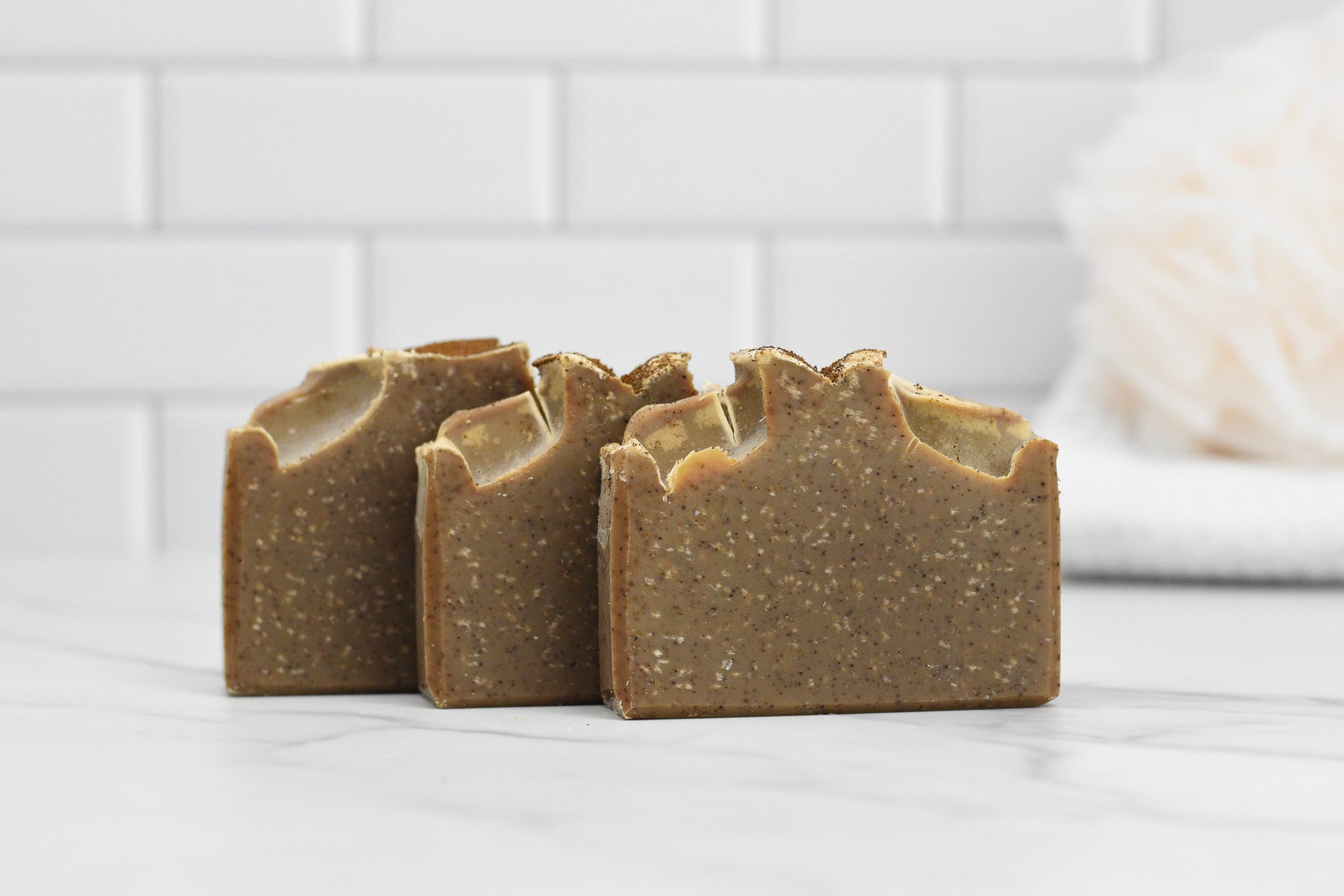 Three bars of 570 Soap Co.'s Coffee Scrub Bar Soap: Wood Shelf Cafe Coffee are displayed on a white marble surface against a backdrop of white tiles. A fluffy white bath sponge is slightly blurred in the background, and the soap's exfoliating kaolin clay promises a refreshing cleanse.