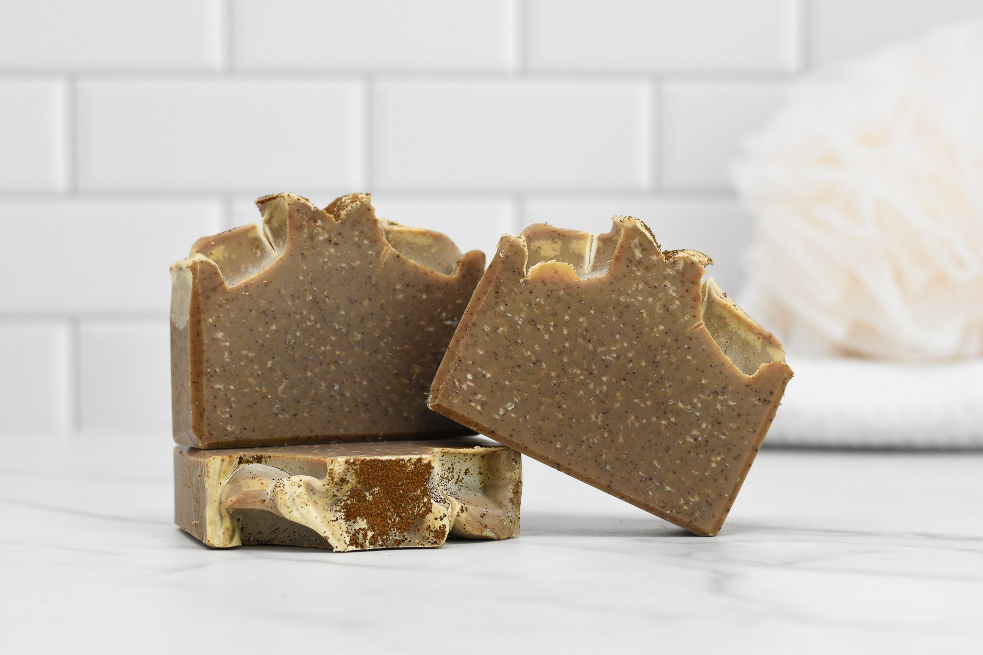 Three Coffee Scrub Bar Soaps from 570 Soap Co., with a speckled texture, are stacked and arranged on a white marble surface. Infused with exfoliating kaolin clay, they promise a gentle scrub. The background features white subway tiles and a blurred bath sponge.