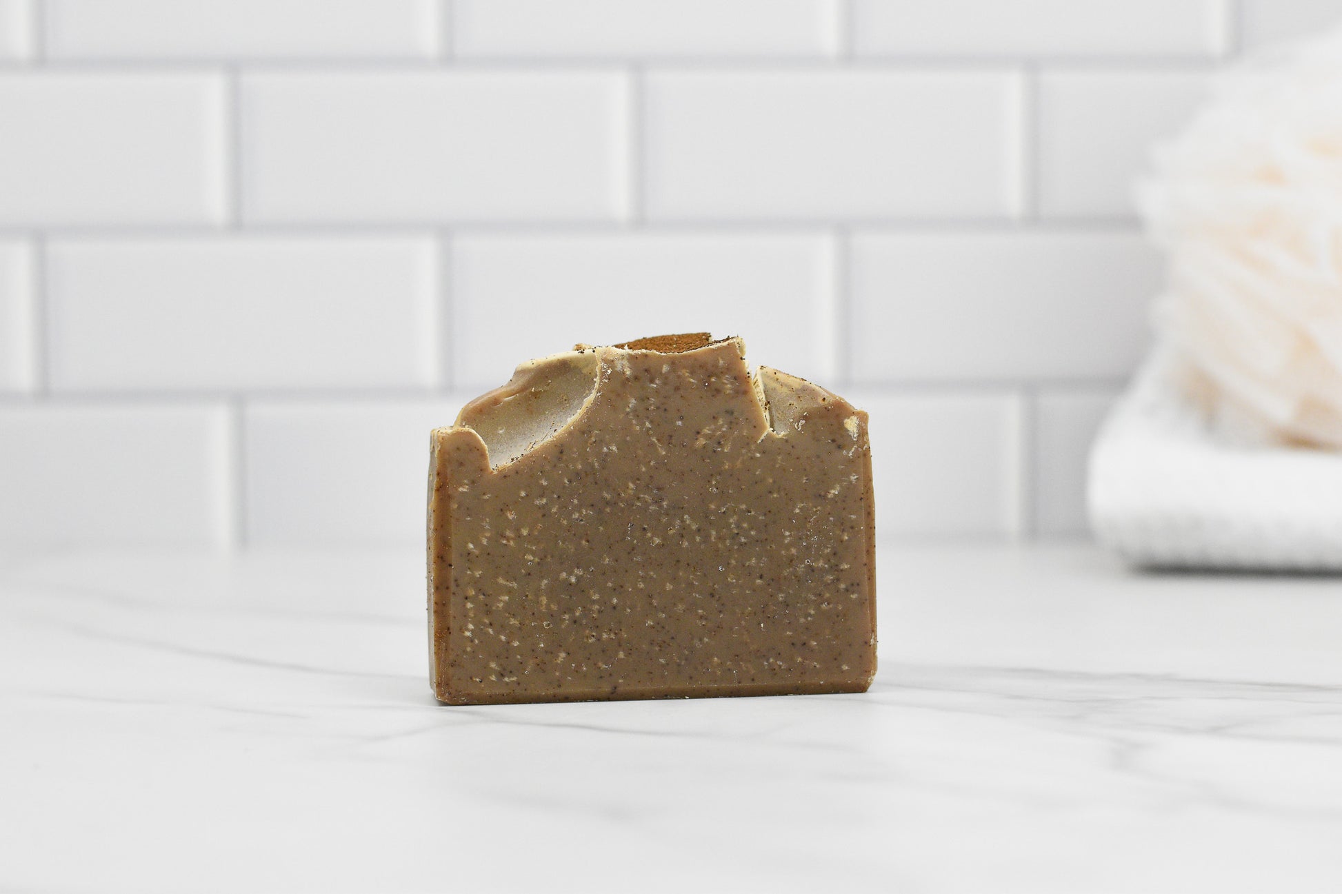 A bar of Coffee Scrub Bar Soap: Wood Shelf Cafe Coffee by 570 Soap Co., enriched with exfoliating kaolin clay, sits on a marble countertop. The background features a wall of white subway tiles, and to the right, a softly blurred loofah adds to the ambiance.