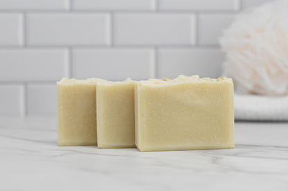 Three bars of 570 Soap Co.'s Oatmeal Bar Soap - All Natural are arranged on a white marble surface. The scene is set against a white tiled wall with the addition of a soft, white loofah, creating a clean and minimalist aesthetic.