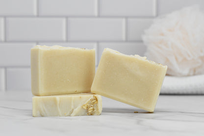 Stacked on a white marble surface against a tiled backdrop are three bars of 570 Soap Co.'s Oatmeal Bar Soap - All Natural. These oatmeal soap bars feature an enriching touch of olive oil, while a softly blurred white bath sponge in the background completes the serene and pure ambiance.