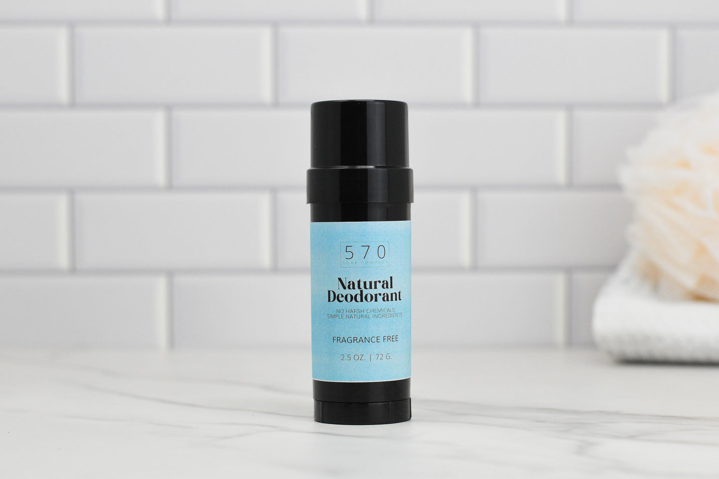 A black stick of natural deodorant with a light blue label sits on a marble surface. The label reads 570 Soap Co., Natural Deodorant - Essential Oils - Stay Fresh Naturally - VEGAN Formula. Featuring natural ingredients for effective odor control, the background showcases white subway tiles and a blurred bath sponge on the right.