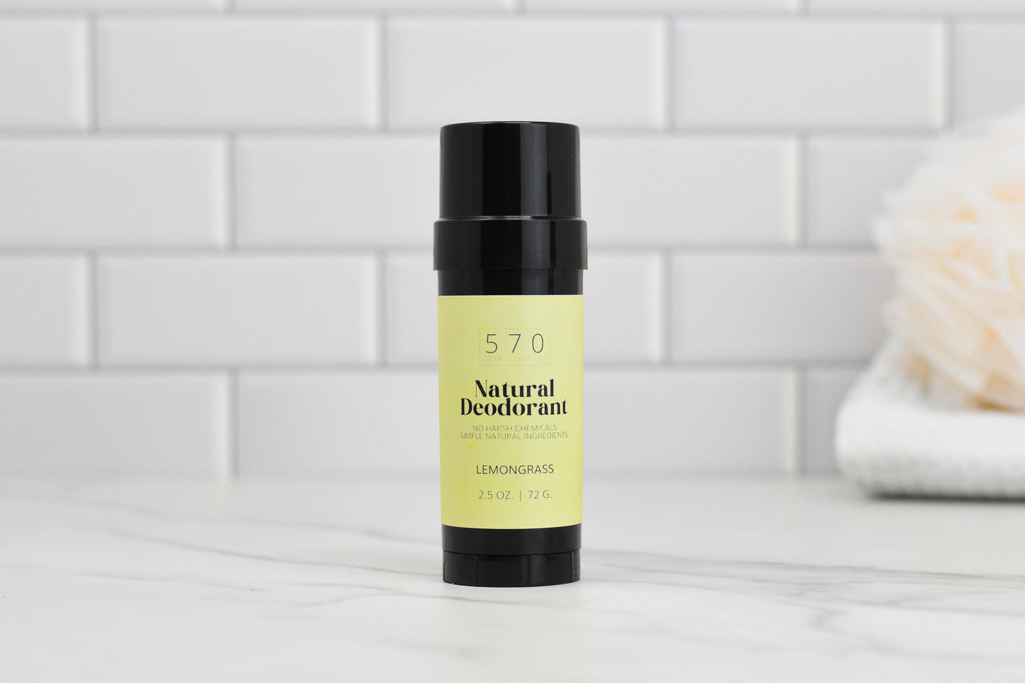 A black cylindrical stick of natural deodorant from 570 Soap Co., formulated with essential oils and a VEGAN composition, has a yellow-green label that reads "Natural Deodorant - Essential Oils - Stay Fresh Naturally." It is set against a white tiled background, with a blurred white towel and light pink object in the distance.