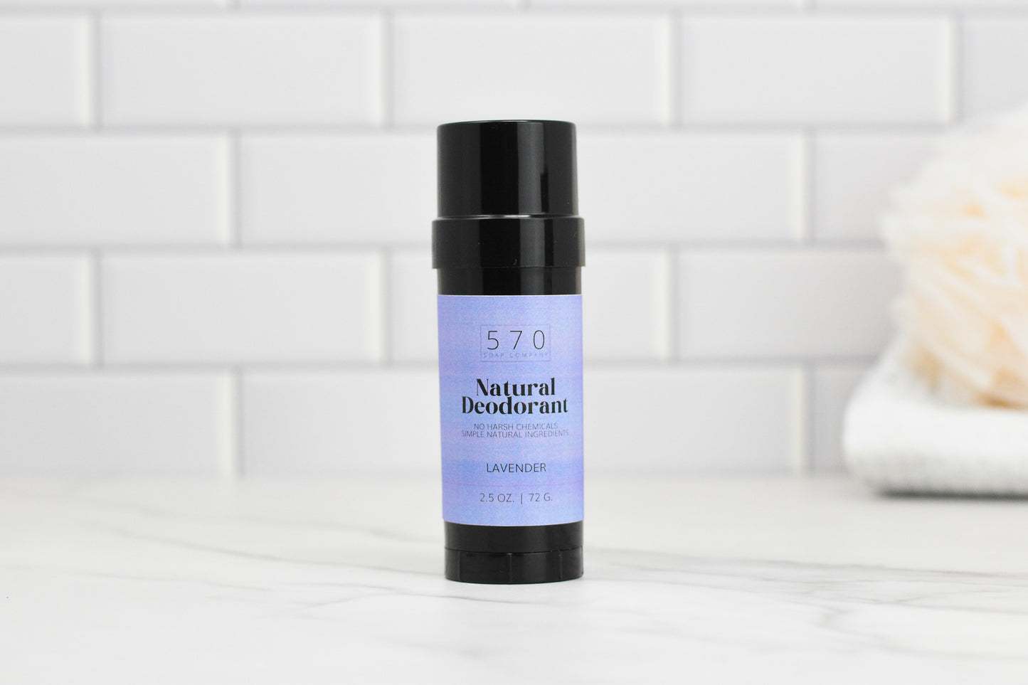 A stick of the Natural Deodorant - Essential Oils from 570 Soap Co., known for its odor control, is placed on a marble surface. The black container displays a light purple label with "570 Soap Co." written on it. In the background, a white towel is visible against a white tiled wall.