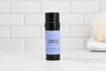 A stick of the Natural Deodorant - Essential Oils from 570 Soap Co., known for its odor control, is placed on a marble surface. The black container displays a light purple label with "570 Soap Co." written on it. In the background, a white towel is visible against a white tiled wall.