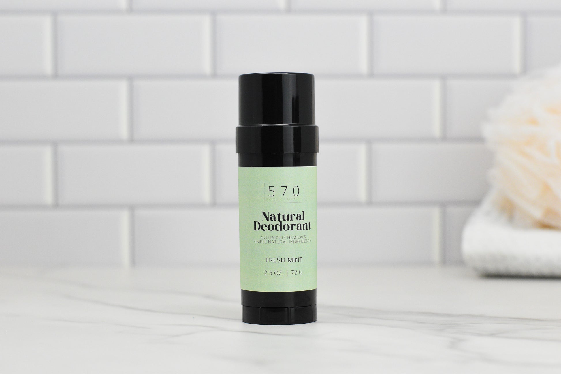 A black tube of 570 Soap Co.'s Natural Deodorant - Essential Oils - Stay Fresh Naturally, featuring a Fresh Mint scent and odor control made with natural ingredients, rests elegantly on a marble countertop. The background displays white subway tiles and a softly blurred bath sponge.