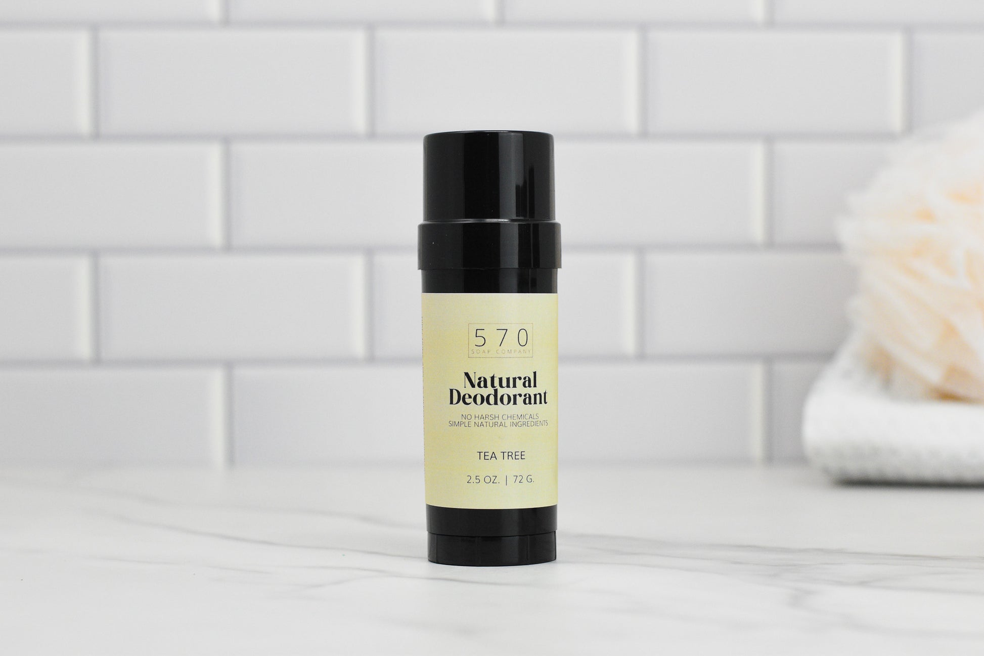 A black cylindrical stick of 570 Soap Co.'s Natural Deodorant, featuring a VEGAN formula with essential oils for effective odor control, sits elegantly on a white marble surface. The background showcases white subway tiles, with a soft towel partially visible on the right.