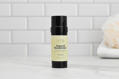 A black cylindrical stick of 570 Soap Co.'s Natural Deodorant, featuring a VEGAN formula with essential oils for effective odor control, sits elegantly on a white marble surface. The background showcases white subway tiles, with a soft towel partially visible on the right.