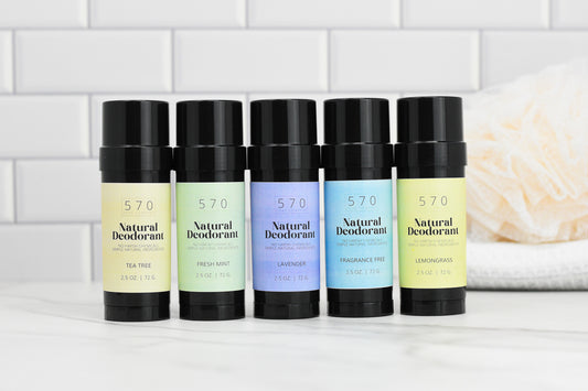 Five black sticks of Natural Deodorant from 570 Soap Co. with vibrant labels, featuring effective odor control and scents such as Tea Tree, Fresh Mint, Lavender, Fragrance-Free, and Lemongrass—all made from natural ingredients—are aligned on a white marble surface. White tiles can be seen in the background.