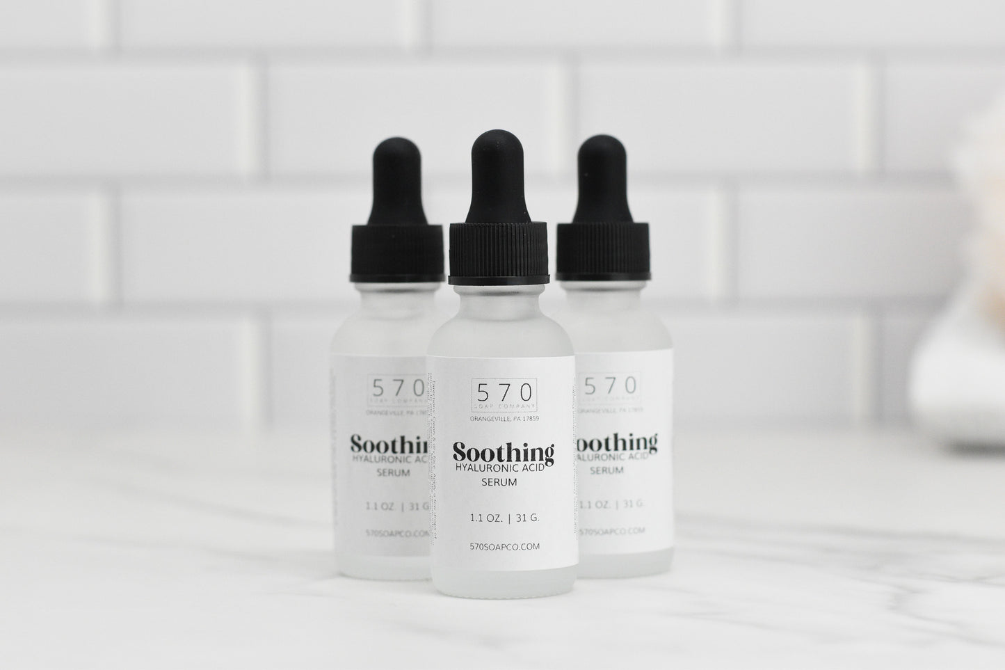 Three bottles of 570 Soap Co.'s Soothing Hyaluronic Acid Serum, enriched with Vitamin B5, are arranged with black dropper tops on a white marble surface. The backdrop of white subway tiles complements the clean and minimalist setting, ideal for hydrating and calming your skin.