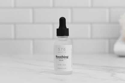 A bottle of 570 Soap Co.'s Soothing Hyaluronic Acid Serum with a dropper cap sits elegantly on a marble surface, ready to hydrate and calm your skin. In the background, white subway tiles enhance its sleek appearance.