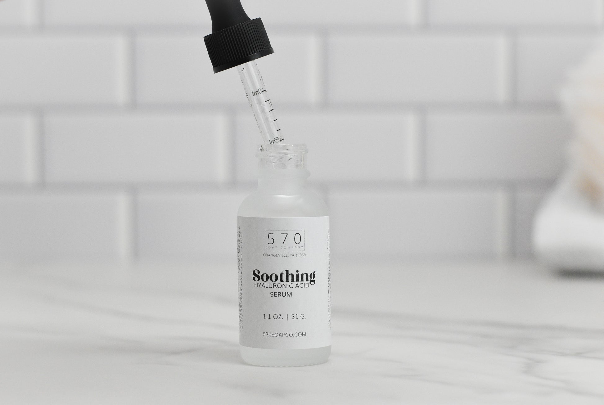 A dropper is poised above a small glass bottle labeled with the name Soothing Hyaluronic Acid Serum by 570 Soap Co., featuring a capacity of 1 fl oz (31 g) and infused with Vitamin B5 to hydrate and soothe your skin. The bottle rests on a marble surface in front of a white tiled backdrop.