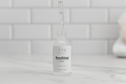 A dropper delicately dispenses liquid into a clear glass bottle labeled "Soothing Hyaluronic Acid Serum by 570 Soap Co." on a marble surface. The backdrop of white subway tiles enhances the sense of purity as the serum hydrates and soothes your skin.